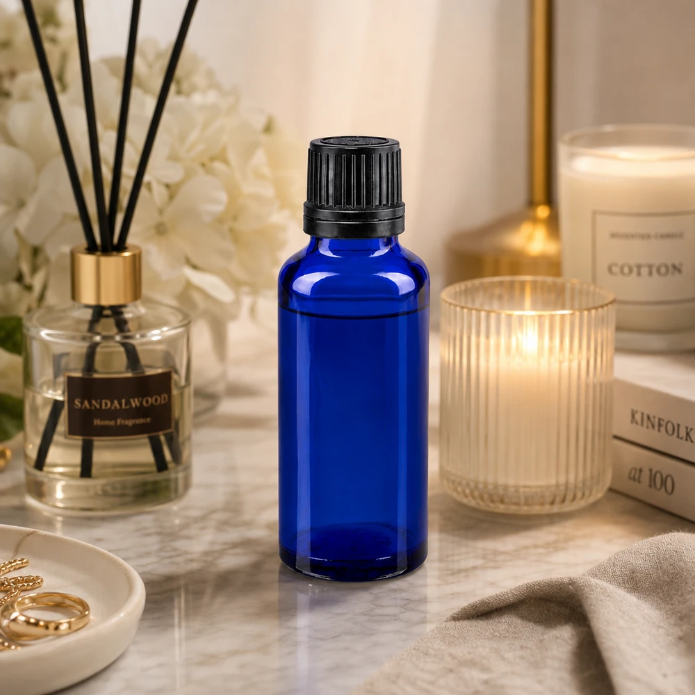 Bottiglia da 30ml blu (Glo.) GR OV nero/8mm ApoGlas Flacone di vetro blu da 30 ml con tappo contagocce nero su una superficie di marmo, accanto a una candela profumata e a un diffusore di profumo per ambienti