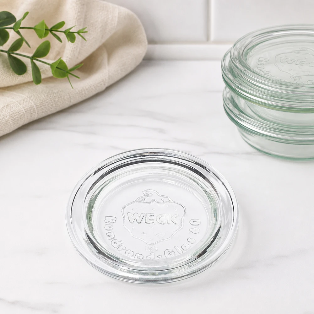 Coperchio in vetro WECK RR60 vista impilata di diversi coperchi sovrapposti su marmo con telo da cucina e ramo verde