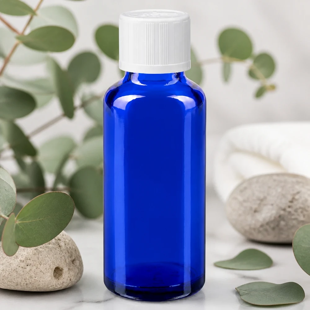 Contagocce blu da 30 ml. STD KiSi bianco/1,2mm ApoGlas Flacone da farmacia da 30 ml blu cobalto con tappo KiSi bianco su fondo marmorizzato, tra rami di eucalipto