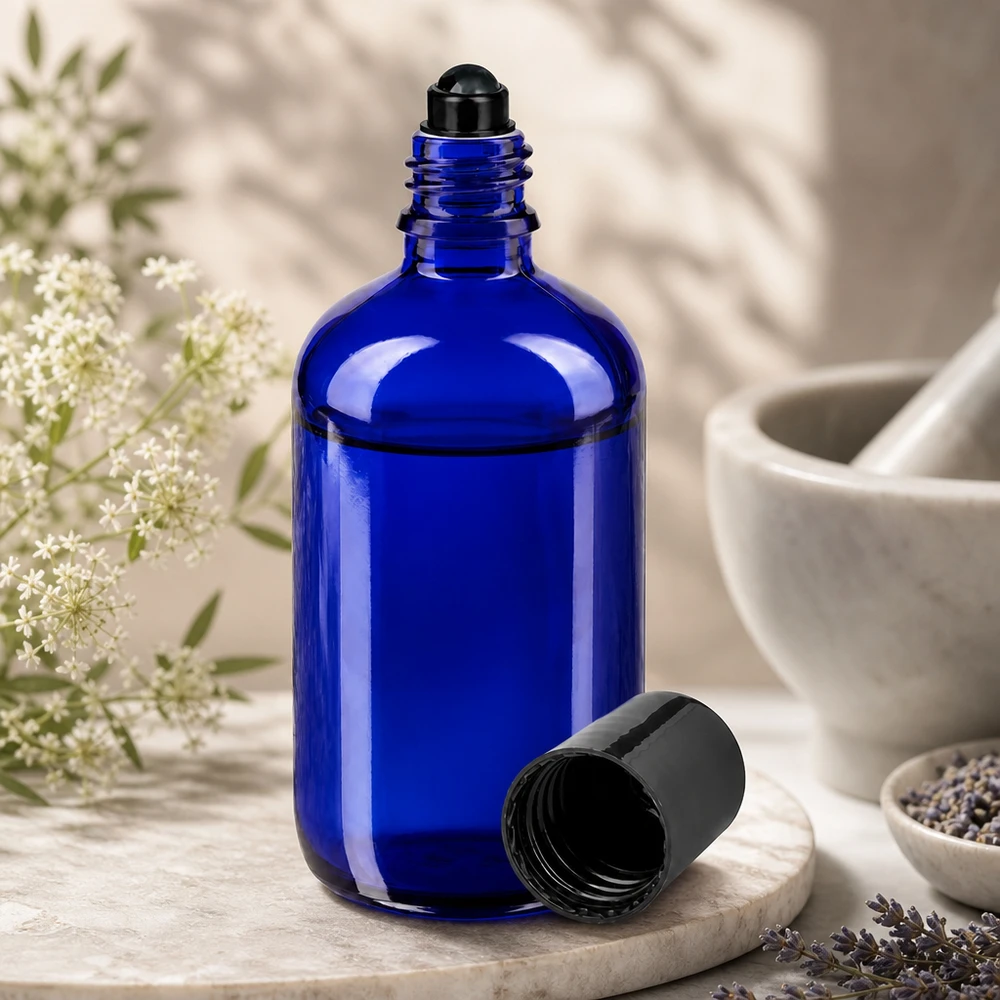 Flacone roll-on 100ml blu STD nero ApoGlas Flacone di vetro blu cobalto da 100 ml con applicatore a sfera nero, aperto, accanto a fiori di lavanda e a un mortaio