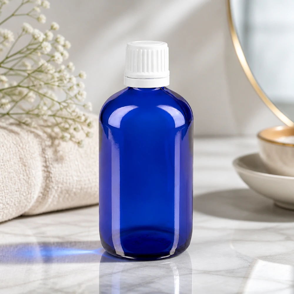 Bottiglia di vetro blu da 100 ml con tappo OV bianco su una superficie di marmo, accanto a un asciugamano e a fiori secchi