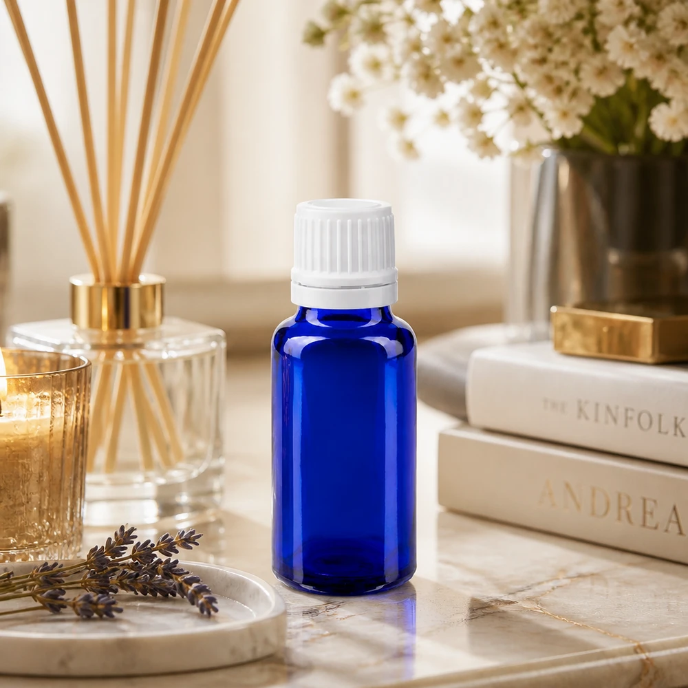 Bottiglia da farmacia blu da 20ml. SV OV bianco/11mm ApoGlas Flacone di vetro blu da 20 ml con tappo a scatto bianco su un tavolino di marmo accanto a un diffusore di profumo per ambienti