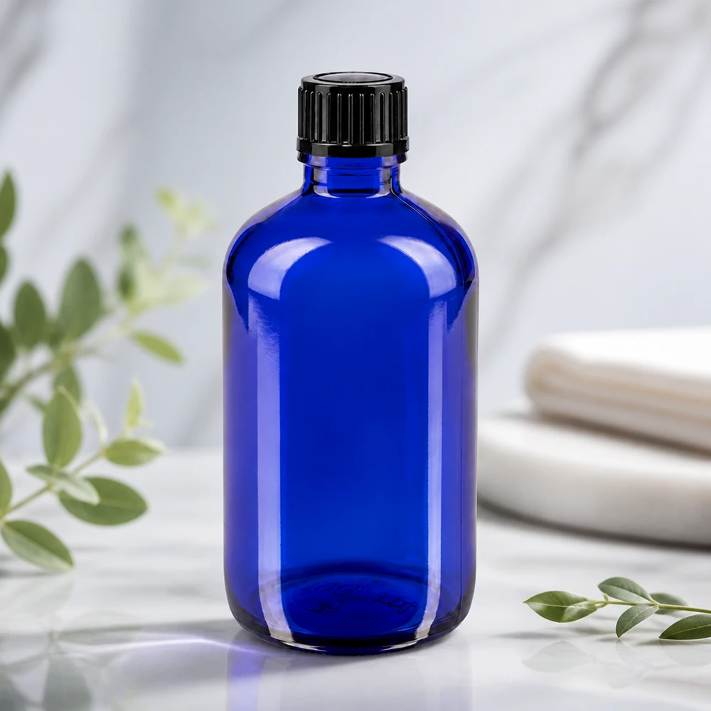 Flacone contagocce blu da 100 ml STD nero/1mm ApoGlas Bottiglia di vetro blu da 100 ml con tappo nero a coste su una superficie di marmo bianco con ramoscelli di eucalipto
