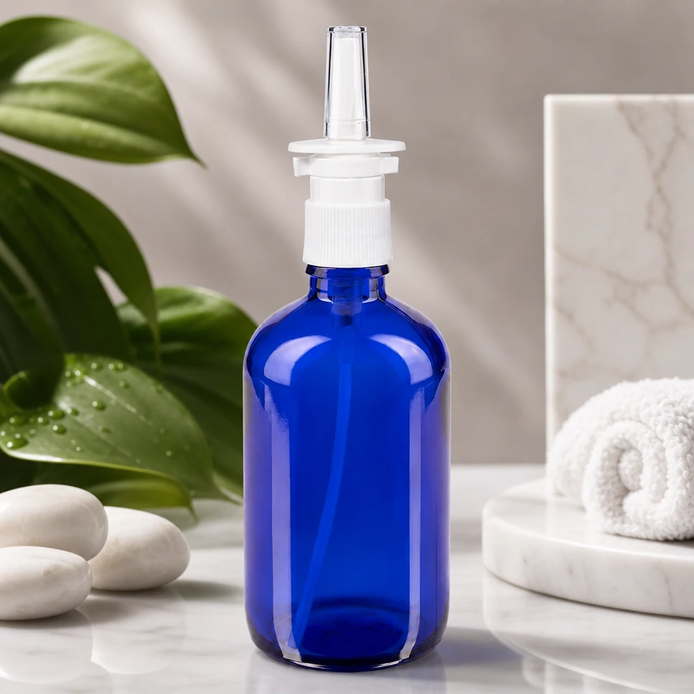 Flacone di vetro da 100 ml blu cobalto con beccuccio spray, appoggiato su marmo bianco accanto a pietre per il benessere
