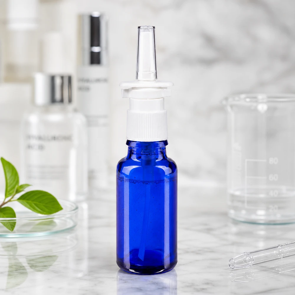Flacone spray nasale da 20 ml in vetro da farmacia blu con nebulizzatore bianco, accanto a un becher e all'acido ialuronico