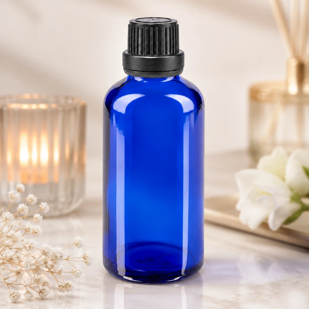 Flacone di vetro da 50 ml blu cobalto con tappo a vite nero e contagocce, accanto a una candela e a dei fiori