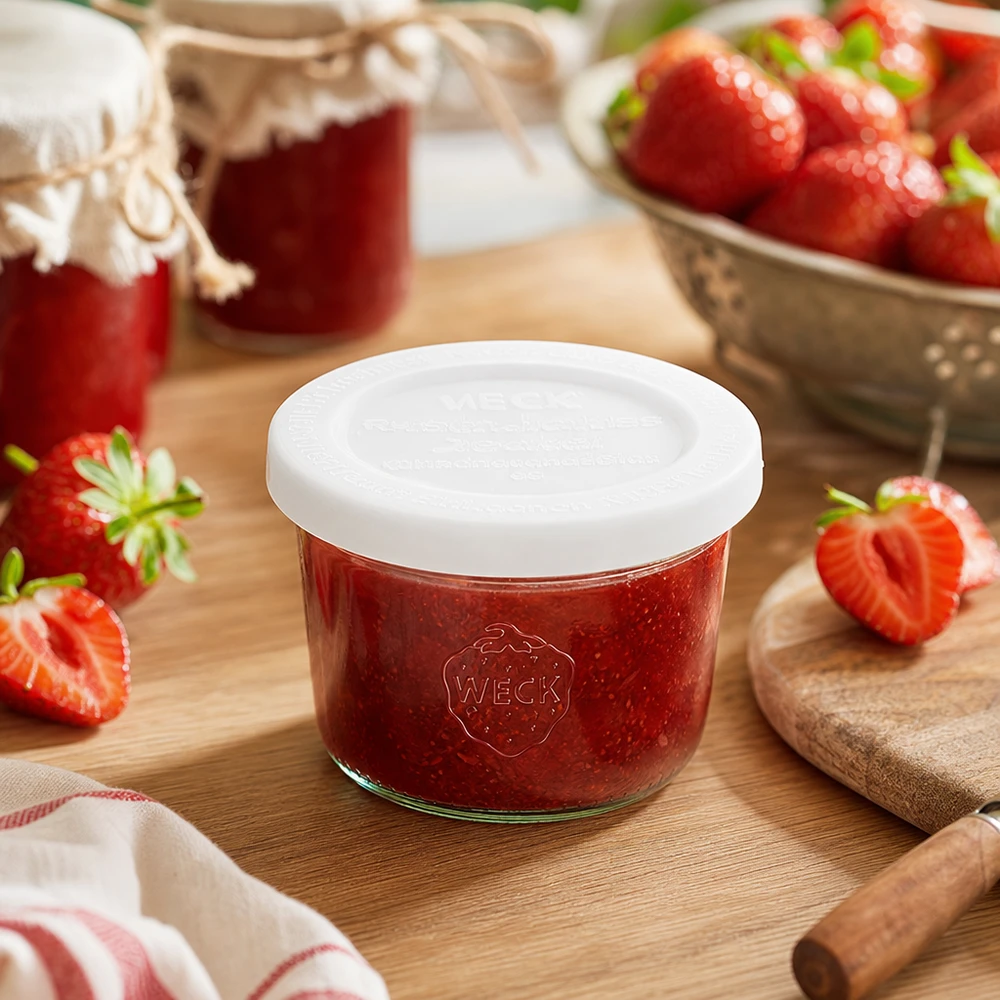 Barattolo da 80 ml con coperchio salva-freschezza WECK RR60 WECK 80ml tumbler RR60 riempiti di marmellata di fragole su una tavola di legno circondata da fragole fresche e barattoli di marmellata