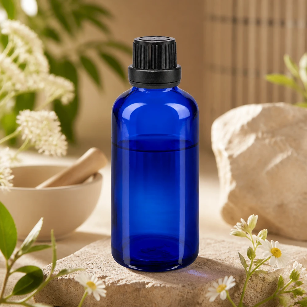 Flacone di vetro da 50 ml blu cobalto con tappo a vite nero, appoggiato su una pietra naturale con fiori di camomilla e un mortaio
