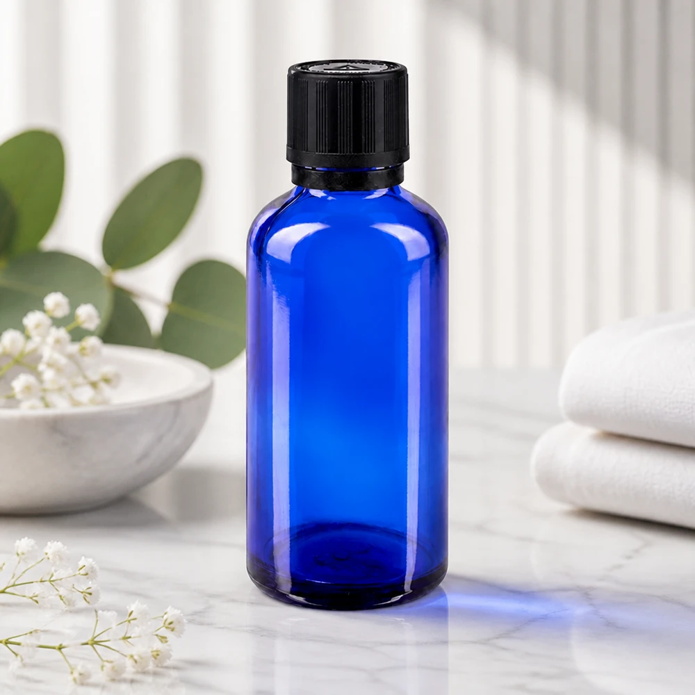 Contagocce blu da 50 ml. OV KiSi Bli nero/1mm ApoGlas Flacone di vetro blu da 50 ml con tappo a vite scanalato su una superficie di marmo, accanto a fiori bianchi di gipsofila